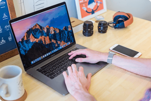 Person using a laptop on a wooden desk with an iPhone, headphones, camera lenses, and a coffee mug nearby