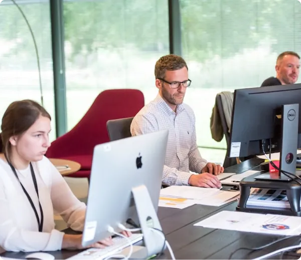 Focused man working at a desk with multiple computer screens in a bright office environment with green outdoor views
