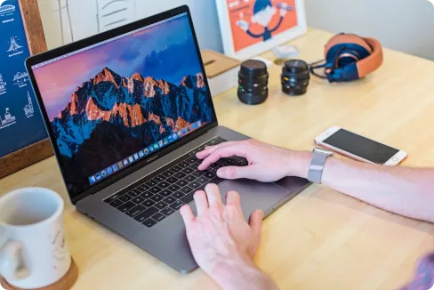 Person using a laptop on a wooden desk with an iPhone, headphones, camera lenses, and a coffee mug nearby