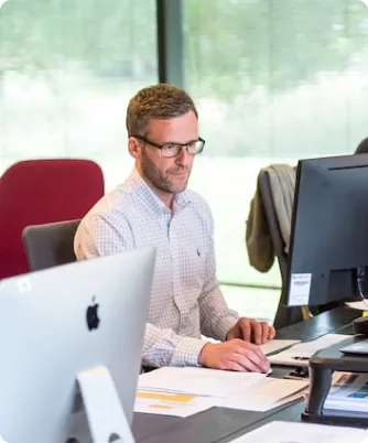 Team of professionals working diligently at their desks with computers in an office with large windows offering natural light and a view of greenery
